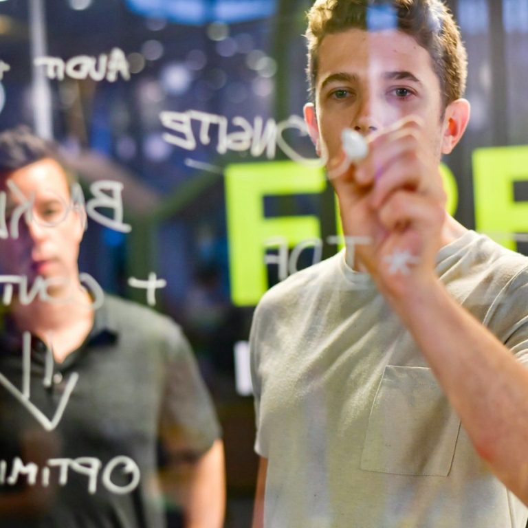 Zwei junge Männer stehen an einer Glasscheibe und schreiben mit einem Marker.